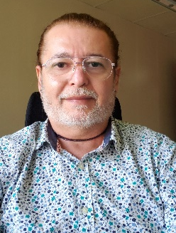 Homem branco, cabelos castanho escuro, possui barba grisalha, olhos castanhos, está usando óculos transparente e está com uma camisa nas cores branco e azul.
